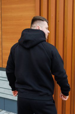 Tarz sahibi siyah kapüşonlu bir genç adam arkası kameraya dönük duruyor ve modern ahşap bir arka plana karşı gururla şehir modasını sergiliyor.