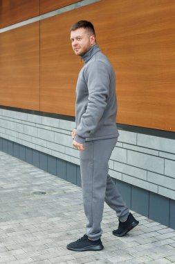 Genç bir adam modaya uygun gri spor kıyafeti sergiliyor, çağdaş mimarinin arka planına güvenle karşı çıkıyor, yaşam tarzı ve moda çekimleri için mükemmel..