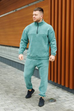 Şık bir genç adam, kendine güvenen bir şekilde şık bir deniz mavisi eşofman modeli, spor için ideal, günlük gezintiler ya da rahatlama, cazip bir şehir zeminine karşı kurulmuş.