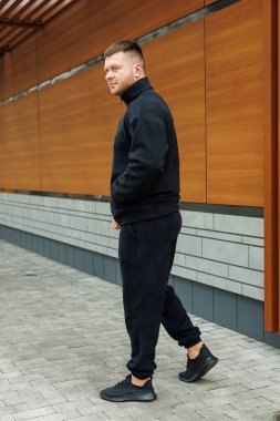 Gösterişli siyah eşofman giymiş şık bir genç adam, şehir tarzı ve konforunu sergileyerek, güvenli bir şekilde dışarıda yürür, hepsi gösterişli modern mimarinin arka planına karşıdır.