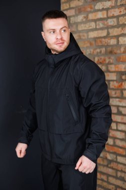 Parlak siyah ceketli, kendine güvenen, atletik bir adam, desenli tuğla bir duvarın önünde poz veriyor, spor modası ve pazarlama için güç ve stili somutlaştırıyor.