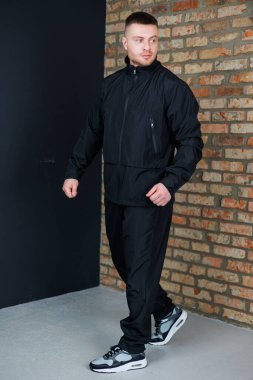 Kendine güvenen bir adam modern bir ortamda şık ve şık bir siyah spor kıyafeti sergilemekten gurur duyar. Endüstriyel tuğla duvarın olduğu bir ortamda yürüyor.