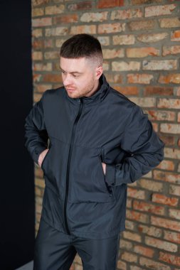 Modern siyah kıyafetli genç bir adam, elleri ceplerinde, desenli tuğladan bir duvara yaslanmış duruyor. Moda ve yaşam tarzı temalarını sergilemek için mükemmel.