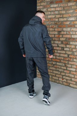 Şık bir insan, şık bir kapüşonlu ceket ve modaya uygun koşucular giyerek, bir tuğla duvarın yanında poz veriyor, kentsel atletizm tarzını ve benzersiz moda anlayışını gözler önüne seriyor.