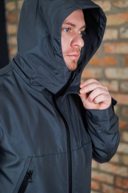 Yakın plan, modaya uygun kapüşonlu ceketini ayarlayan bir adamı, taşralı tuğla bir duvara karşı modern şehir modasını sergiliyor, yaşam tarzı ve moda markaları için mükemmel.