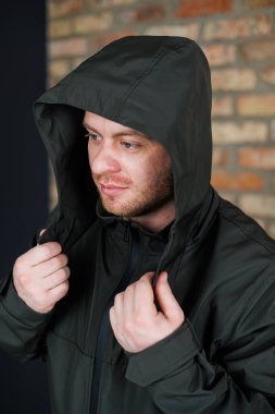 Koyu renk kapüşonlu bir ceket giymiş genç bir adam ellerini yakasına koyup düşünceli bir şekilde poz veriyor. Duvara yaslanmış, karakter katıyor.