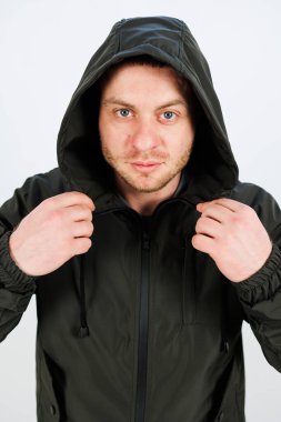 Kararlı ve odaklanmış bir ifadeye sahip genç bir adam şık kapüşonlu ceketini düzeltiyor, kendine güvenerek hem stil hem de duruşunu minimalist bir ortamda sergiliyor