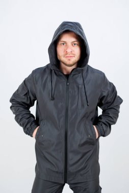 Siyah kapüşonlu ceket giyen şık bir erkek manken, elleri kalçalarının üzerinde, beyaz bir arka plana yaslanmış, modern sokak kıyafetlerini sergiliyor..