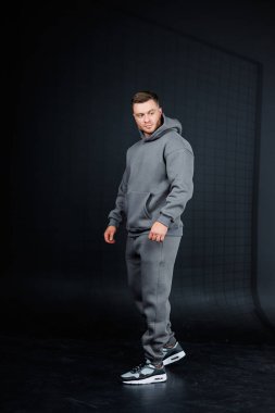 Kendine güvenen genç bir adam şık gri kapüşonlu bir poz veriyor ve modaya uygun koşucuları karanlık bir arka plana itiyor. Günümüz modern spor giyim tarzının özünü somutlaştırıyor.