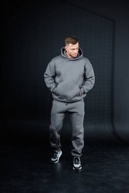 Şık bir genç adam rahat gri bir kapüşonlu ve rahat bir eşofmanla poz veriyor, çağdaş şehir modasını davetkar minimalist bir stüdyo ortamında sergiliyor.