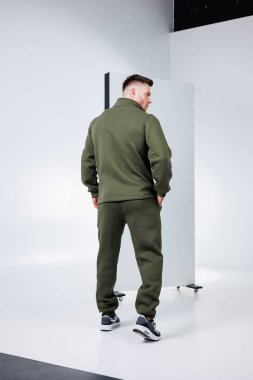 Son moda zeytin rengi eşofmanlı bir erkek model minimalist bir arka plana karşı duruyor, modern spor tarzını göstererek konforu zarafetle birleştiriyor.