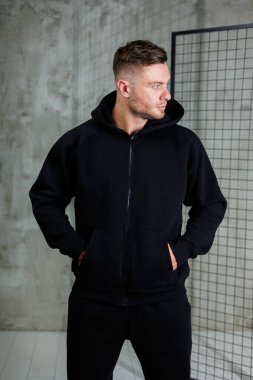 Modayı takip eden genç bir adam, şık, siyah fermuarlı kapüşonlu bir kapüşonlu giyinerek, görünüşünü ön plana çıkaran minimalist kentsel bir arka plana karşı modern gündelik moda sergiliyor.
