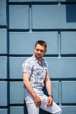 Moda desenli gömleği ve parlak beyaz pantolonu olan şık bir adam canlı mavi bir duvara karşı duruyor. Modern tarzını yaz güneşinin altında sergiliyor.