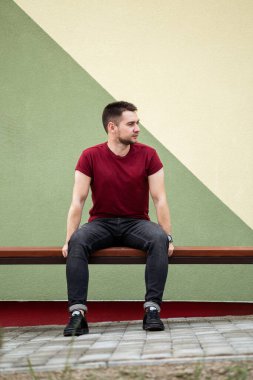 Şık bir genç adam modern ahşap bir bankta rahat eder, canlı kırmızı bir tişört ve mavi kot pantolon giyer, renkli geometrik bir arka plan ile tasasız titreşimleri arttırır.