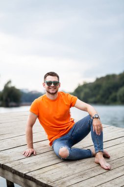 Parlak turuncu tişörtlü ve kot pantolonlu şık bir genç sakin bir gölün kenarındaki sade bir rıhtımda dinleniyor, şık bir selfie çekerken güneşin ve esintinin tadını çıkarıyor.