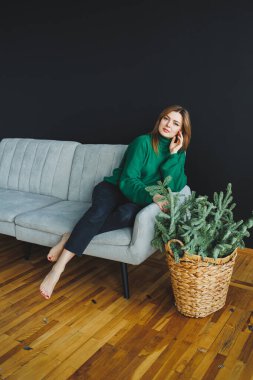 Zarif bir kadın yeşil bir kazakla gri bir kanepeye uzanır. Etrafı canlı iç mekan bitkileriyle çevrilidir. Dekoratif bir sepetle, modern bir ev dekorasyonu estetiğini ön plana çıkarır.
