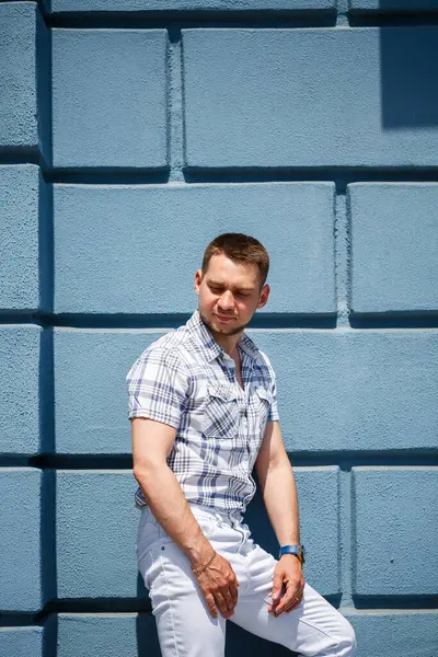 Moda desenli gömleği ve parlak beyaz pantolonu olan şık bir adam canlı mavi bir duvara karşı duruyor. Modern tarzını yaz güneşinin altında sergiliyor.
