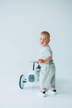 Sevimli kıyafetli çekici bir çocuk, küçük beyaz bir bisiklette neşeyle oynar. Mutlulukla dolu, minimalist bir stüdyoda keyifli anlar yaratır.