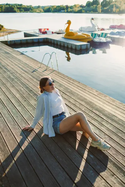 İskelede güzel sarışın bir kadın, beyaz bir gömlek, kısa kot şort ve güneş gözlüğü, nehir kıyısında neşeli bir kadın, deniz kenarında yaz tatili.