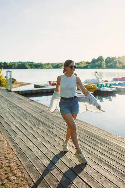Göl kıyısında yürüyen şortlu ve güneş gözlüklü neşeli bir kadın. Kadın eğleniyor ve kavurucu güneşin altında koşuyor. Genç bir kadın göl kenarında yürüyüşe çıkmış..