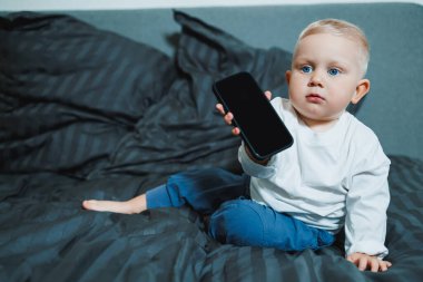 Beyaz tişörtlü ve kot pantolonlu yakışıklı çocuk yatakta oturur ve telefonda çizgi film izler. 2 yaşındaki bir çocuk mobil bir aletle oynuyor. Bebeklerin modern dijital hale getirilmesi.