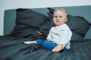 Çocuk telefonu elinde tutuyor. Beyaz tişörtlü, kot pantolonlu yakışıklı çocuk yatakta oturur ve telefonda çizgi film izler. 2 yaşındaki bir çocuk cep telefonuyla oynuyor. Çocukların modern dijitalleştirilmesi.