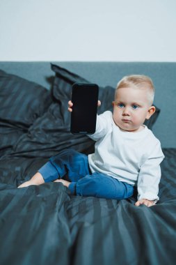 Beyaz tişörtlü ve kot pantolonlu yakışıklı çocuk yatakta oturur ve telefonda çizgi film izler. 2 yaşındaki bir çocuk mobil bir aletle oynuyor. Bebeklerin modern dijital hale getirilmesi.