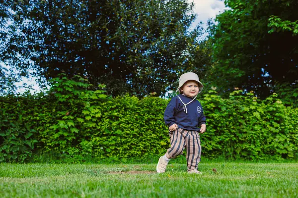 Kafasında panama şapkası olan güzel gülümseyen çocuk şehir parkında yeşil çimlerde koşuyor. Doğada iki yaşında bir çocuğun yürüyüşü. Çocuk doğanın güzelliğinin ve yazın güneşli bir günün tadını çıkarıyor..