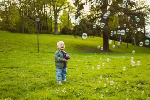 2 yaşında sabun köpükleriyle yeşil çimlerde koşan küçük bir çocuk. Küçük çocuk doğa yürüyüşünün tadını çıkarıyor. Çocuk şehir parkında eğleniyor..