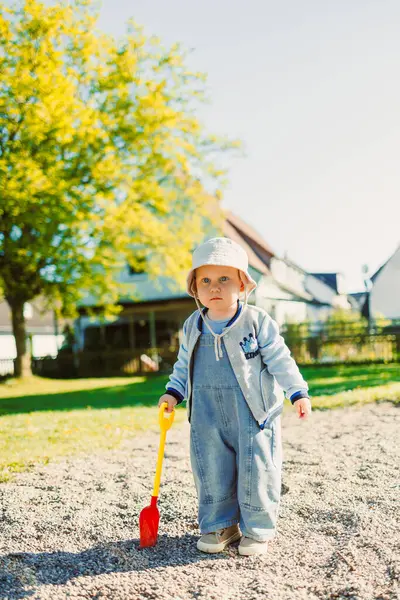 Küçük çocuk oyun parkındaki kum havuzunda kürekle oynuyor. Doğada oynayan bir çocuk. 2 yaşındaki bir çocuk güneşli bir yaz gününde parkta yeşil çimlerin tadını çıkarıyor..
