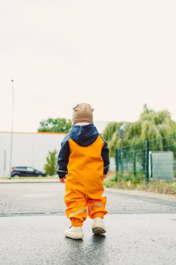 Çamur geçirmez tulumlu ve botları yağmurdan sonra koşan gülen 2 yaşında bir çocuk. Parlak yağmurluklu ve botlu çocuk su birikintilerinde koşuyor. Bulutlu bir günde yürüyüşe çıkmış su geçirmez tulumlu bir çocuk..