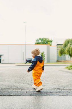 Çamur geçirmez tulumlu ve botları yağmurdan sonra koşan gülen 2 yaşında bir çocuk. Parlak yağmurluklu ve botlu çocuk su birikintilerinde koşuyor. Bulutlu bir günde yürüyüşe çıkmış su geçirmez tulumlu bir çocuk..