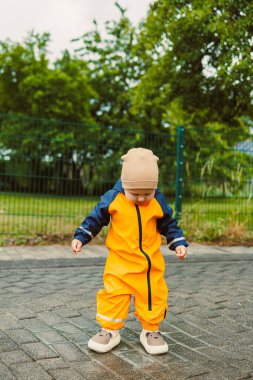 Çamur geçirmez tulumlu ve botları yağmurdan sonra koşan gülen 2 yaşında bir çocuk. Parlak yağmurluklu ve botlu çocuk su birikintilerinde koşuyor. Bulutlu bir günde yürüyüşe çıkmış su geçirmez tulumlu bir çocuk..