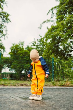 Turuncu yağmurluklu, ıslak yolda su birikintisinde oynayan mutlu 2 yaşında bir çocuk. Çocuk yağmurlu bir günde su geçirmez giysiler ve lastik botlarla. Çocukluk sevinci ve aktif eğlence.