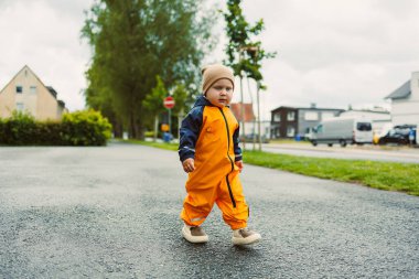 Turuncu yağmurluklu, ıslak yolda su birikintisinde oynayan mutlu 2 yaşında bir çocuk. Çocuk yağmurlu bir günde su geçirmez giysiler ve lastik botlarla. Çocukluk sevinci ve aktif eğlence.