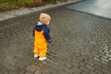 Yağmurlu bir günde asfalt yolda yürüyen sarı tulumlu, 2 yaşında neşeli bir çocuk. Sarı su geçirmez ceketli şık çocuk ve açık havada oynayan lastik botlar..