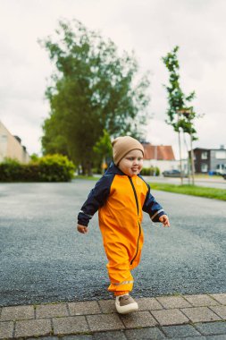 Yağmurlu bir günde asfalt yolda yürüyen sarı tulumlu, 2 yaşında neşeli bir çocuk. Sarı su geçirmez ceketli şık çocuk ve açık havada oynayan lastik botlar..