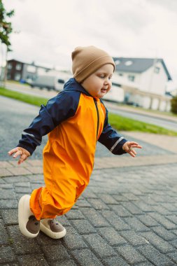 Lastik çizmeli, mavi tulumlu, yağmurlu bir sonbahar gününde parkta ya da bahçede oynayan gülümseyen küçük çocuk.