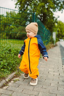 Yağmurlu bir günde asfalt yolda yürüyen sarı tulumlu, 2 yaşında neşeli bir çocuk. Sarı su geçirmez ceketli şık çocuk ve açık havada oynayan lastik botlar..