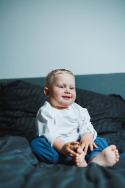 Parlak gülüşü olan, yumuşak yatağında oturan, sıradan kıyafetler giyen, çağdaş bir ev ortamında neşe ve masumiyet gösteren sevimli bir çocuk.