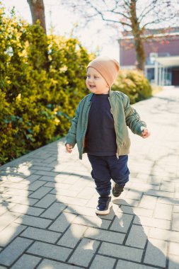 Neşeli bir çocuk parlak ve güneşli bir patika boyunca yürüyor, rahat giysiler içinde neşeli bir gülümseme ile, mutlu yeşil yeşillik ve canlı doğa ile çevrili