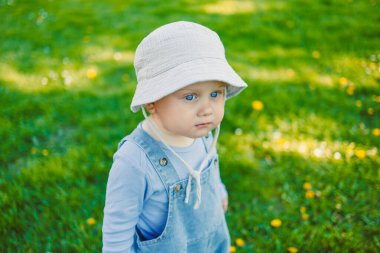 Şirin, hasır şapka ve mavi kot pantolon giyen bir bebek, yemyeşil bir alanda dikiliyor, dışarıda nefis bir günün tadını çıkarıyor, nefes kesici güzelliğin ortasında.