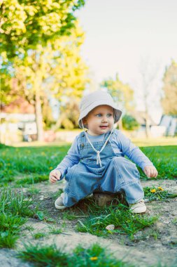 Güneşli bir parkta, neşeli bir çocuk yemyeşil çimenlerin üzerinde oturur, şık bir elbise ve şirin bir şapka giyer, saf neşe ve çocukluğun masumiyetini somutlaştırır.