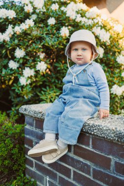 Renkli bir şapka takan neşeli bir çocuk, parlak çiçeklerle çevrili güneşli taştan bir duvarda oturur. Saf çocukluk masumiyetinin ve sonsuz neşenin özünü güzel bir şekilde yakalar.