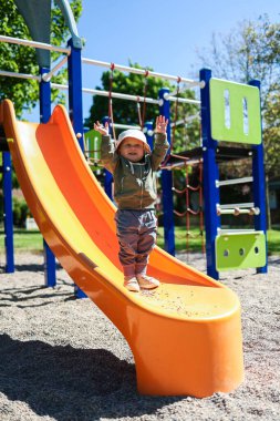 Çok mutlu bir çocuk oyun parkındaki turuncu kaydırağın tepesinde muzaffer bir şekilde durur. Dışarıda sonsuz eğlence ve neşe dolu bir günün tadını çıkarır.