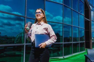 Bir kadın modern bir binanın önünde duruyor, bir elinde dizüstü bilgisayar, diğer elinde dumanı tüten bir fincan kahveyle.