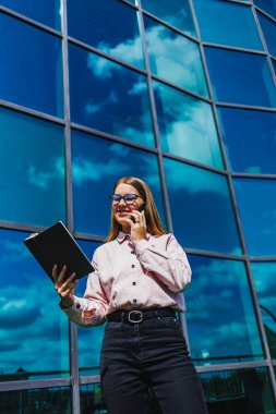 Bir kadın cep telefonuyla konuşurken aynı anda diğer elinde bir tablet tutuyor, açıkça konuşmalarıyla meşgul.