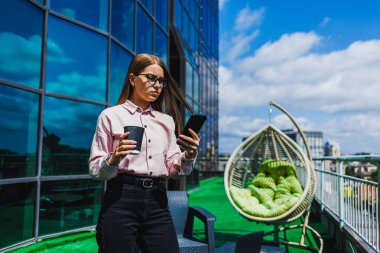Tarz sahibi genç bir kadın güneşli bir balkonda kahvesini yudumlarken tamamen akıllı telefonuna dalmış, modern yaşam tarzında rahatlama ve üretkenliği kusursuz bir şekilde birleştiriyor.