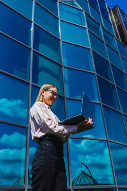 Kendine güvenen bir kadın pozitiflik ve kararlılık sergiliyor, bilgisayarını açık havada çalışırken gülümsüyor, canlı bir şehir ortamında üretkenliği ve gücü somutlaştırıyor.