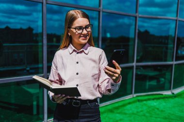 Güneşli bir günde, şık gözlüklü genç bir kadın akıllı telefonunu kontrol ederken ve bir defter tutarken gülümser ve modern iş hayatının dengesini somutlaştırır.